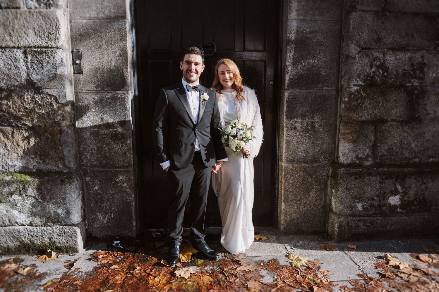 A newlywed couple poses for portraits against a rustic stone wall with autumn leaves.