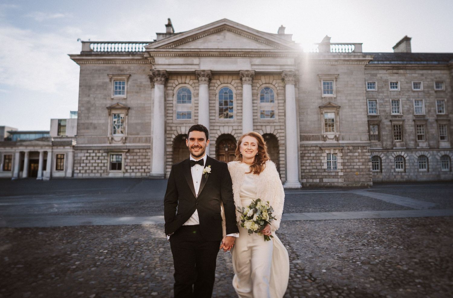 Wedding couple posing in front of grand historic stone building on sunny day.