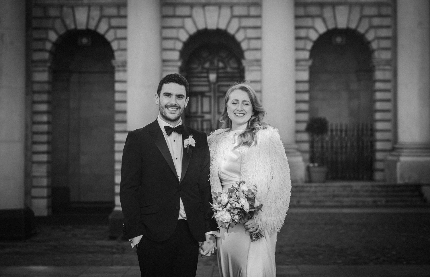 Elegant black and white wedding photo taken in front of a classical stone building with arched windows.