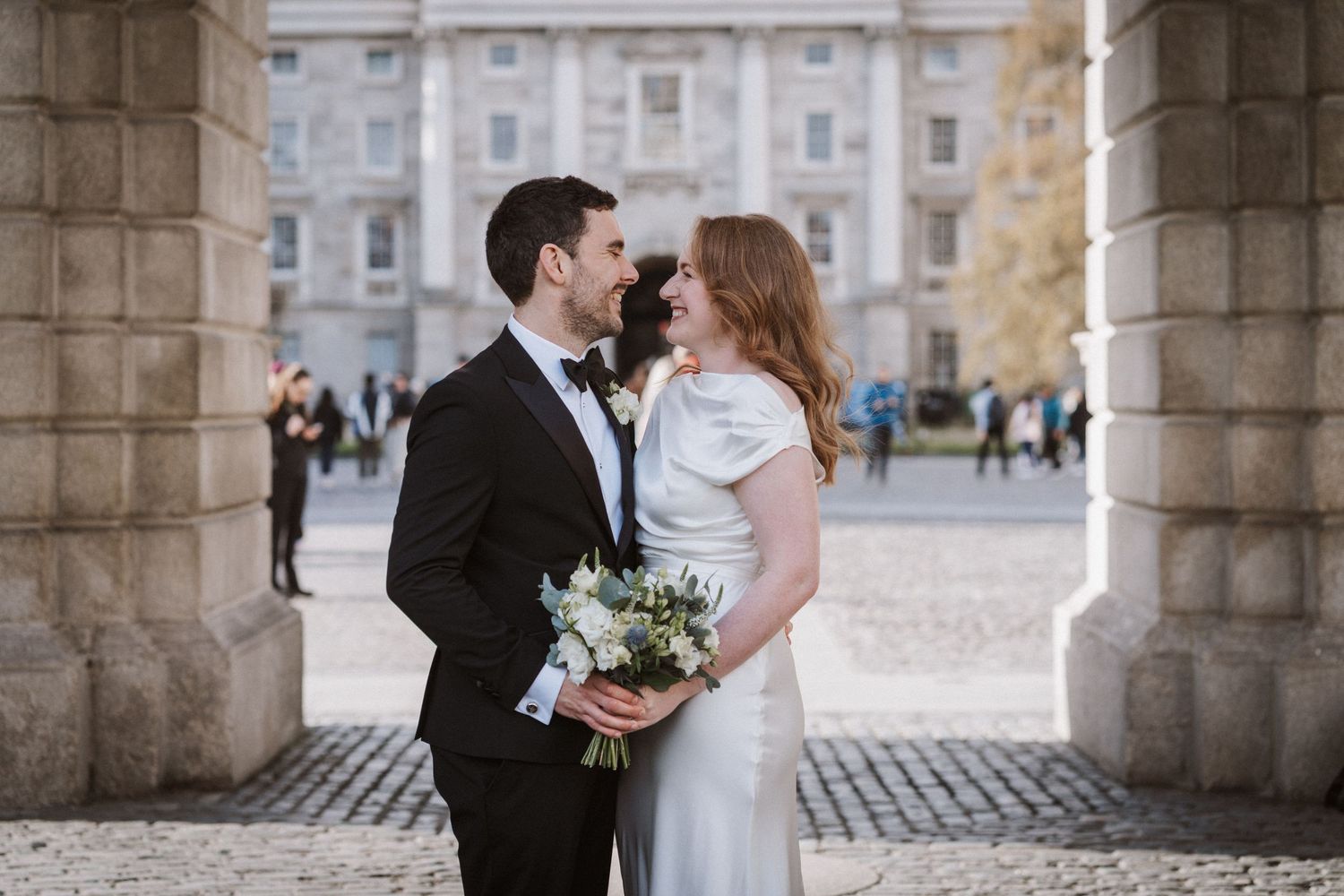 Newlyweds pose under stone archway in urban setting with historic architecture.