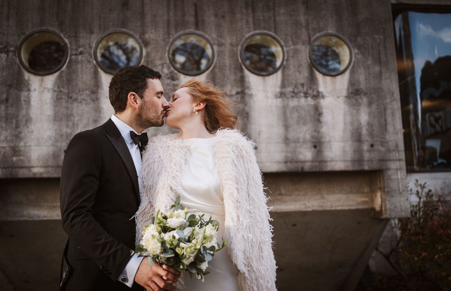 A couple embraces during their wedding celebration against a textured concrete wall backdrop.