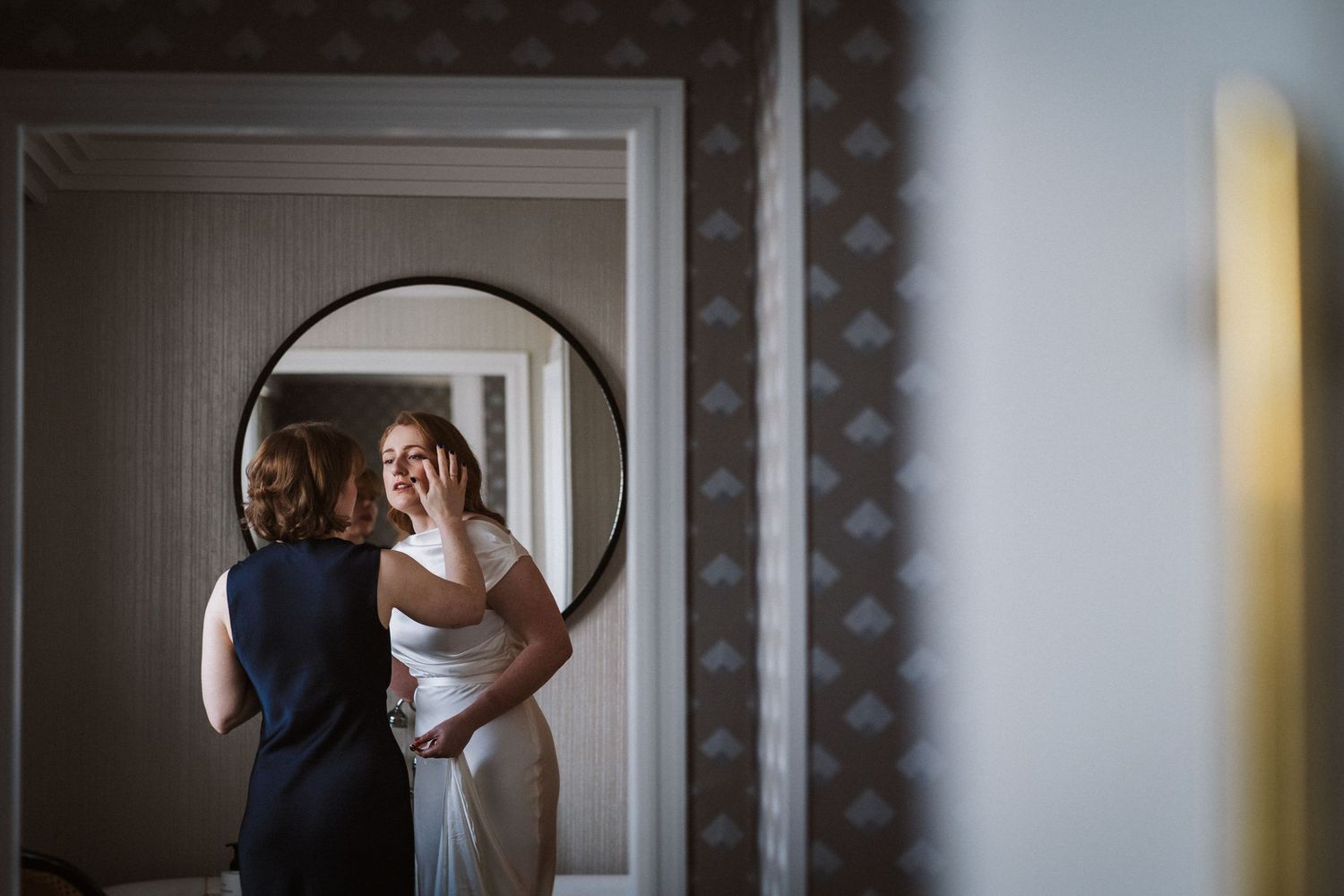 A bride in a white wedding dress adjusts her hair in front of a mirror during wedding preparations.