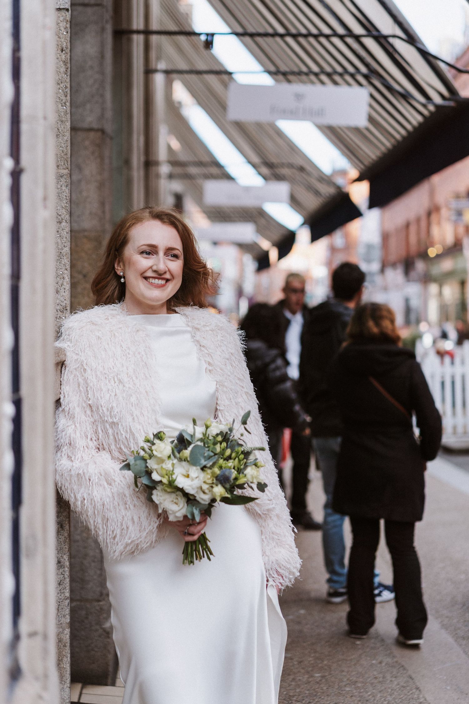 A bride in a white dress with a feathered shawl smiles while holding a winter bouquet on a city street.