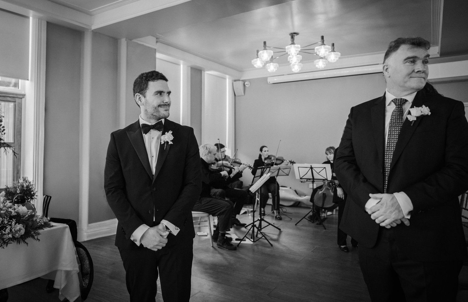 Two men in tuxedos stand in an elegant room during a formal event, captured in black and white photography.
