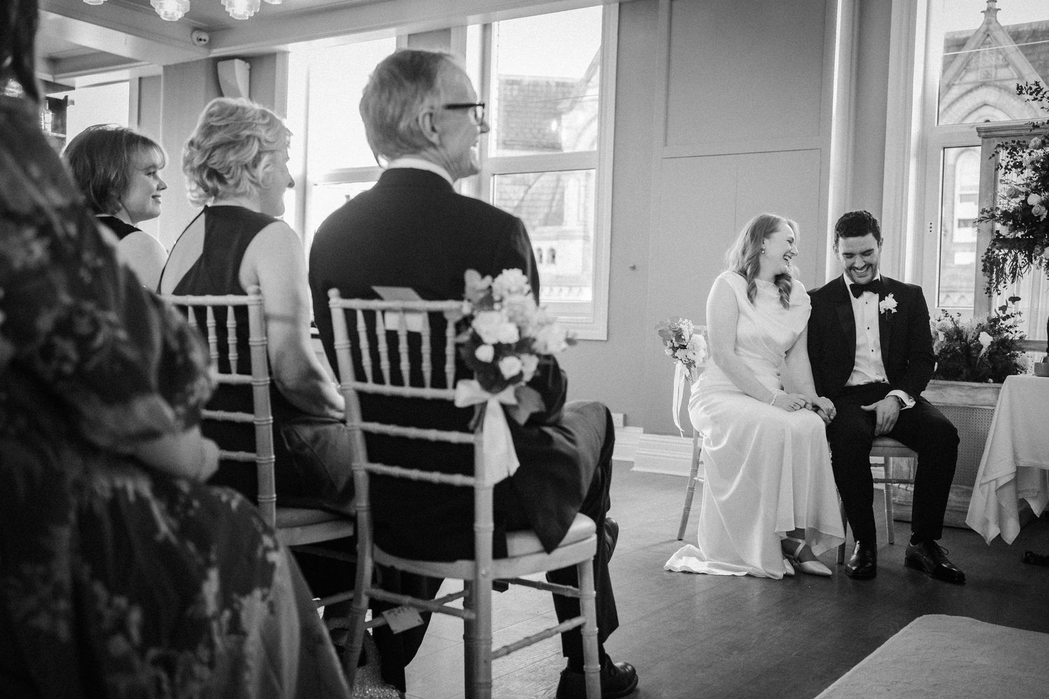 Black and white photo of an intimate wedding ceremony in a bright indoor venue with guests seated in Chiavari chairs.