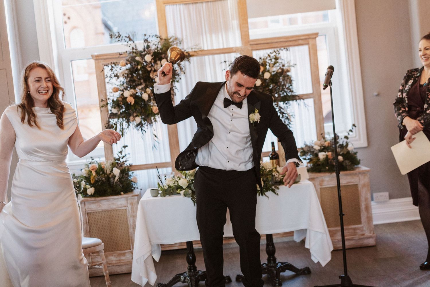 Wedding party celebrates with joyful dancing and laughter in an elegant white-decorated reception venue.