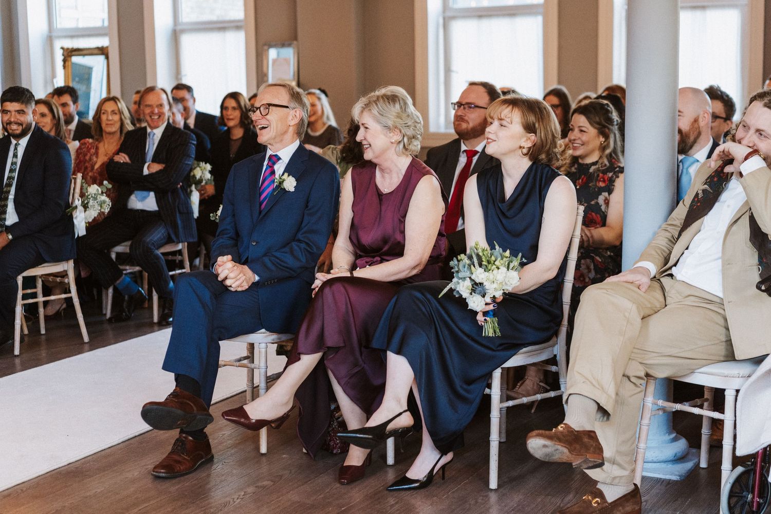 Wedding guests seated in rows during an indoor ceremony with natural lighting through windows.