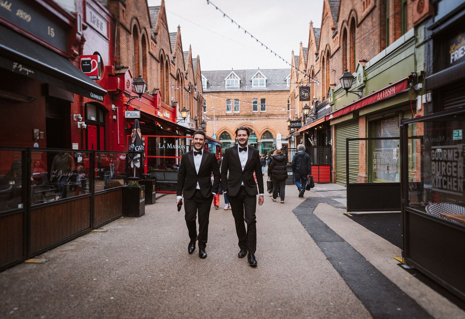 People walking through a historic brick-lined shopping street with storefronts and string lights overhead.
