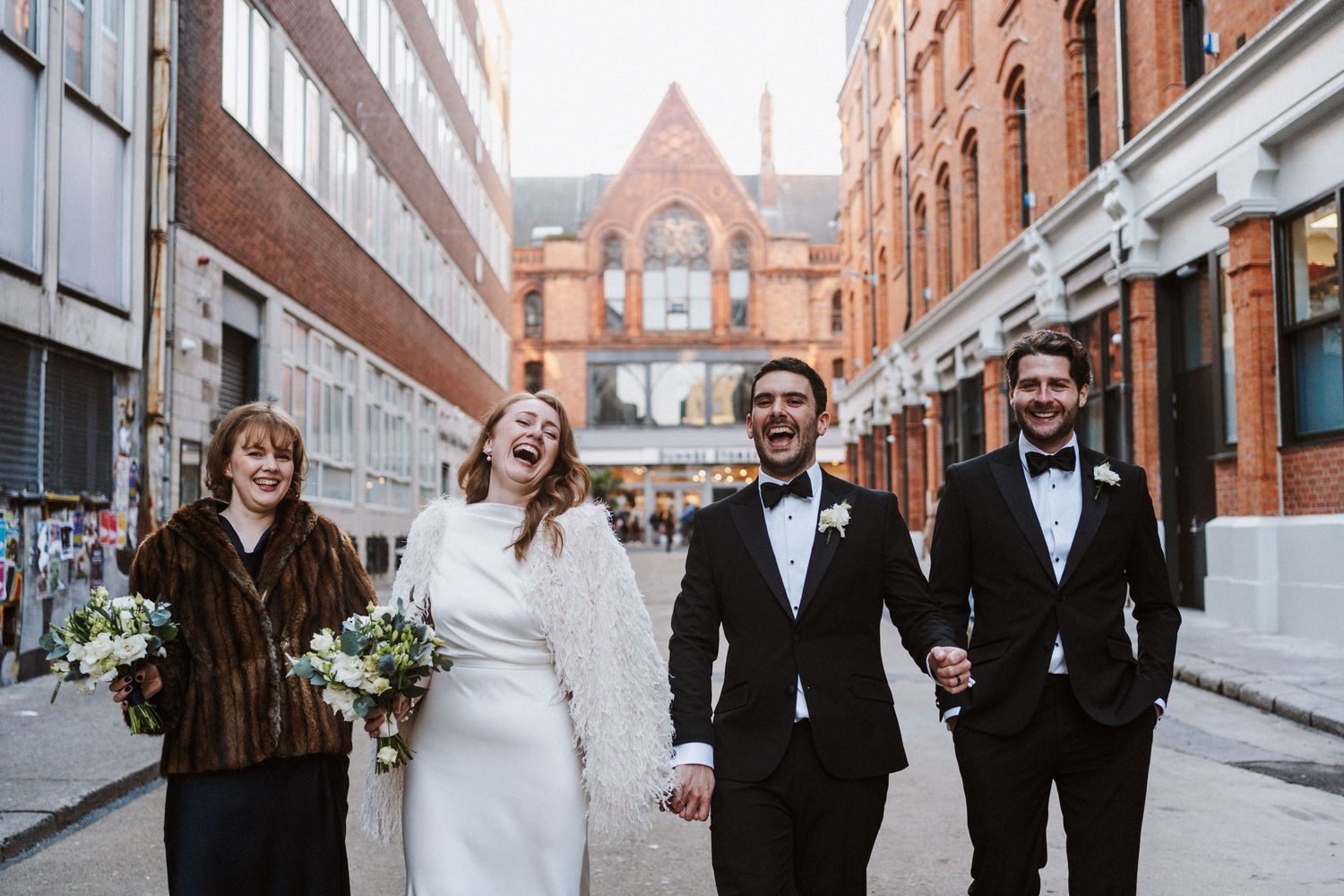 Wedding party walking and laughing together on a brick-lined city street.