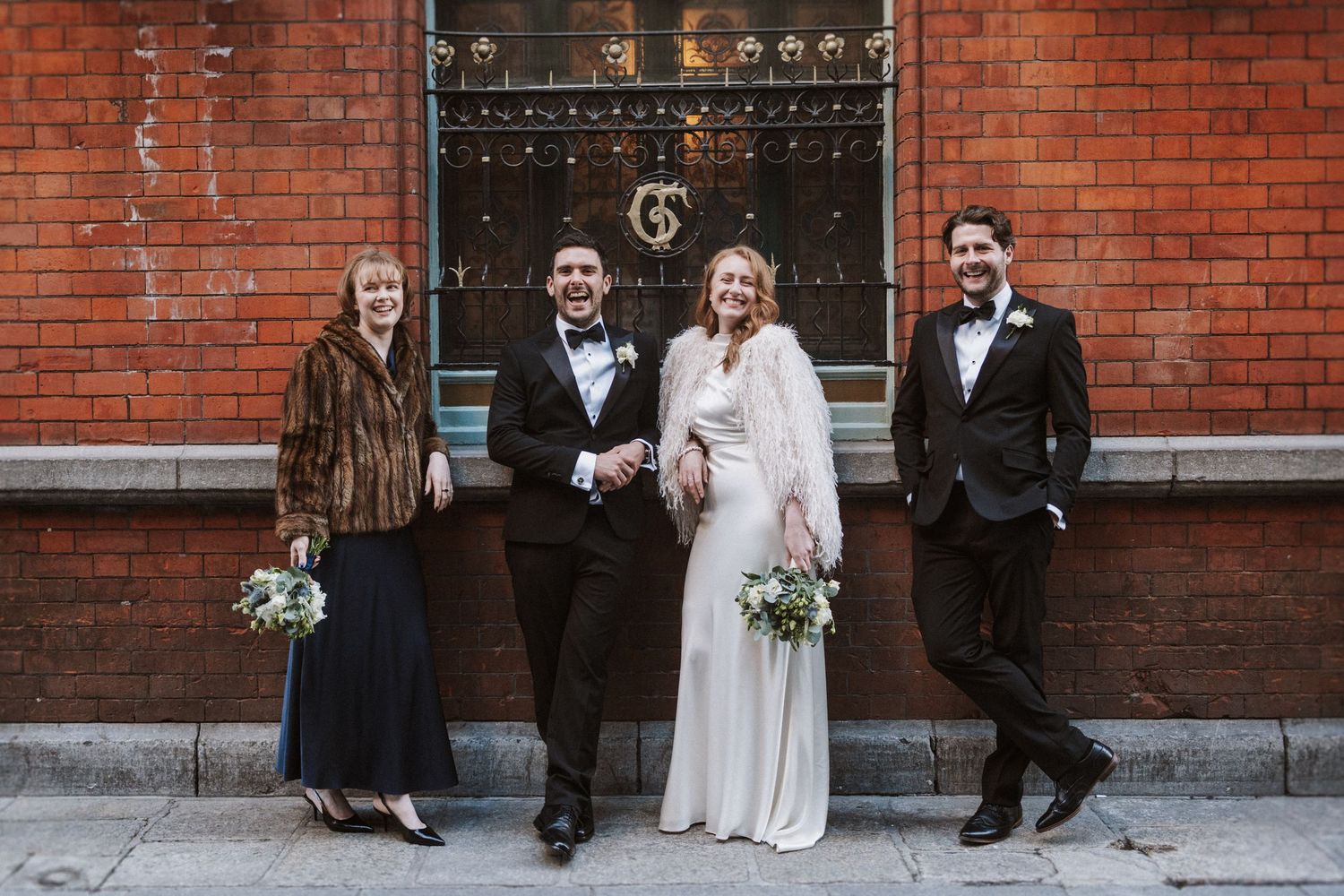Wedding party sitting casually along a rustic brick wall exterior in formal attire.
