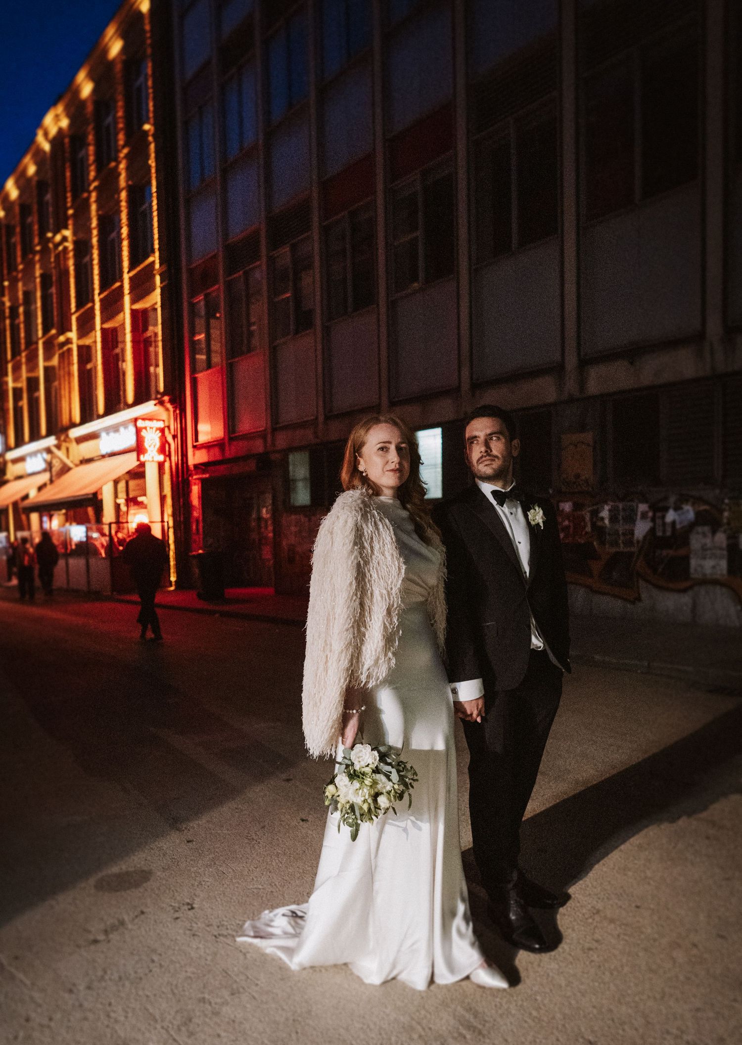 A wedding couple poses for romantic nighttime photos on a city street illuminated by warm glowing lights.
