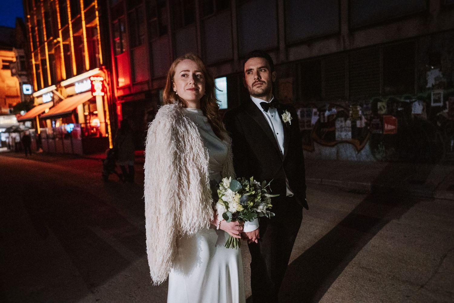 Couple poses for nighttime wedding photos with illuminated urban buildings in the background.
