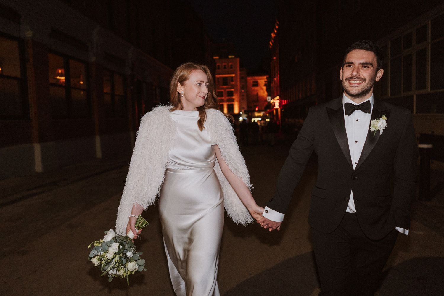 Wedding couple walks hand in hand along dark city street with neon lights glowing in the distance.