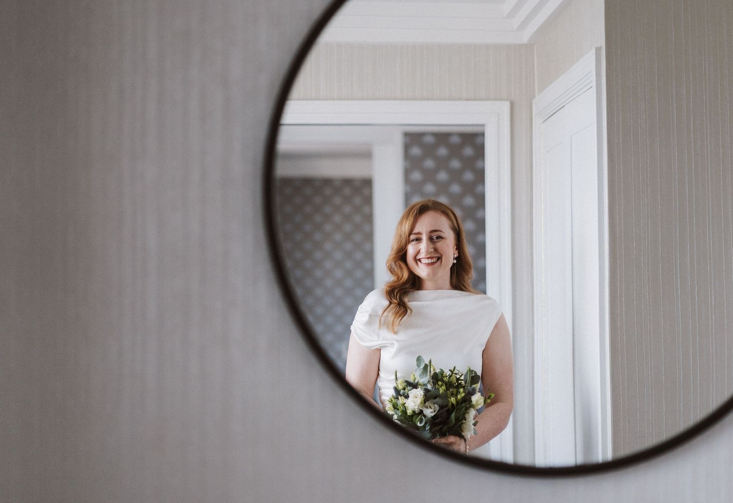 Modern round mirror reflects a bright, minimalist room with textured wallpaper.