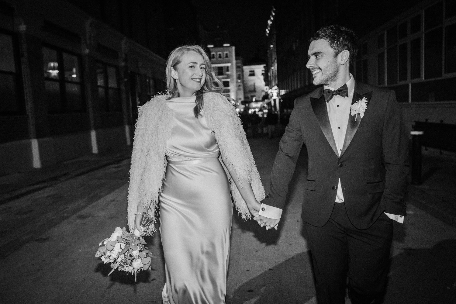 Couple walks hand in hand along a dimly lit urban street at night after their wedding celebration.
