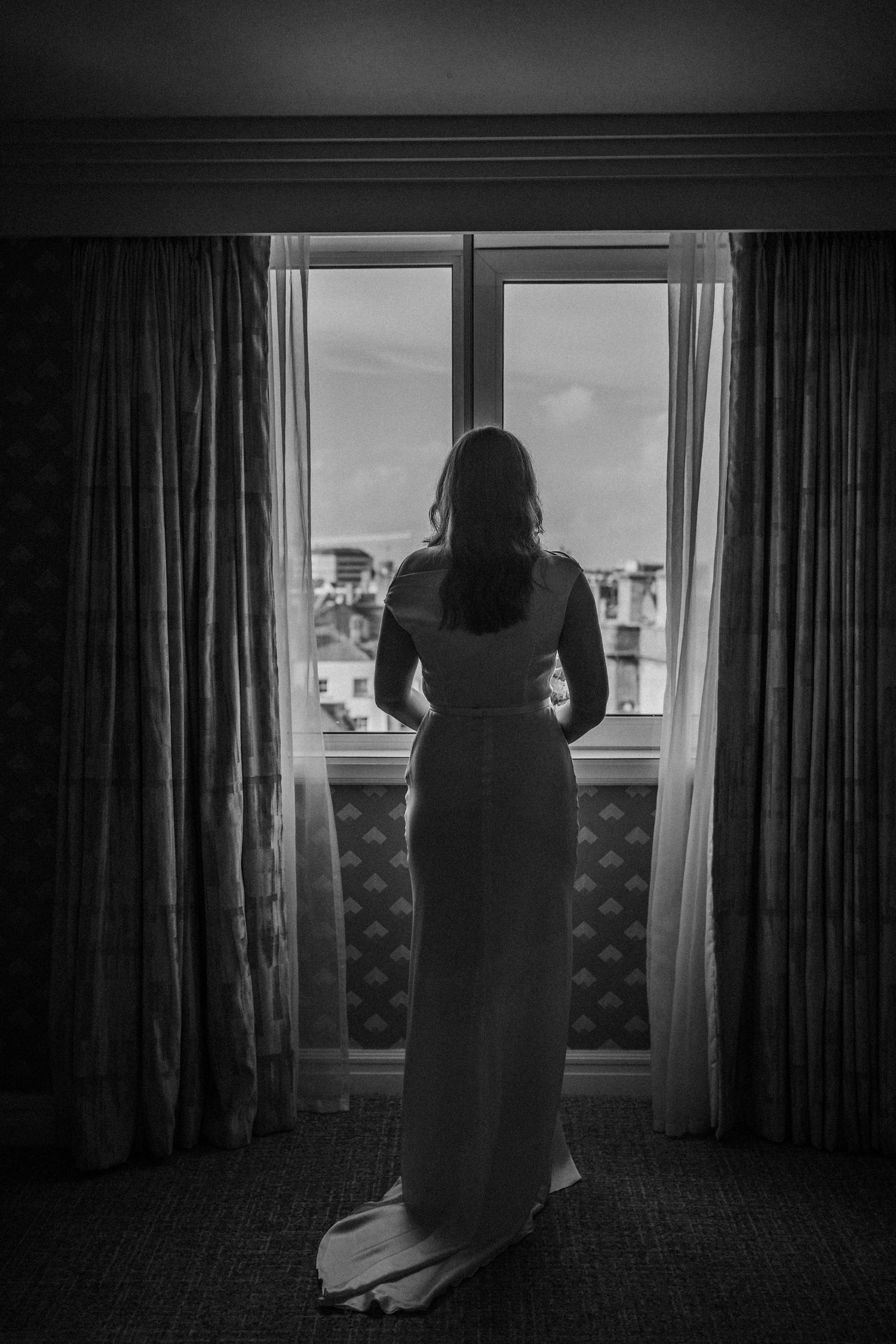 Silhouetted figure stands at window in dark room with curtains creating moody atmospheric scene.