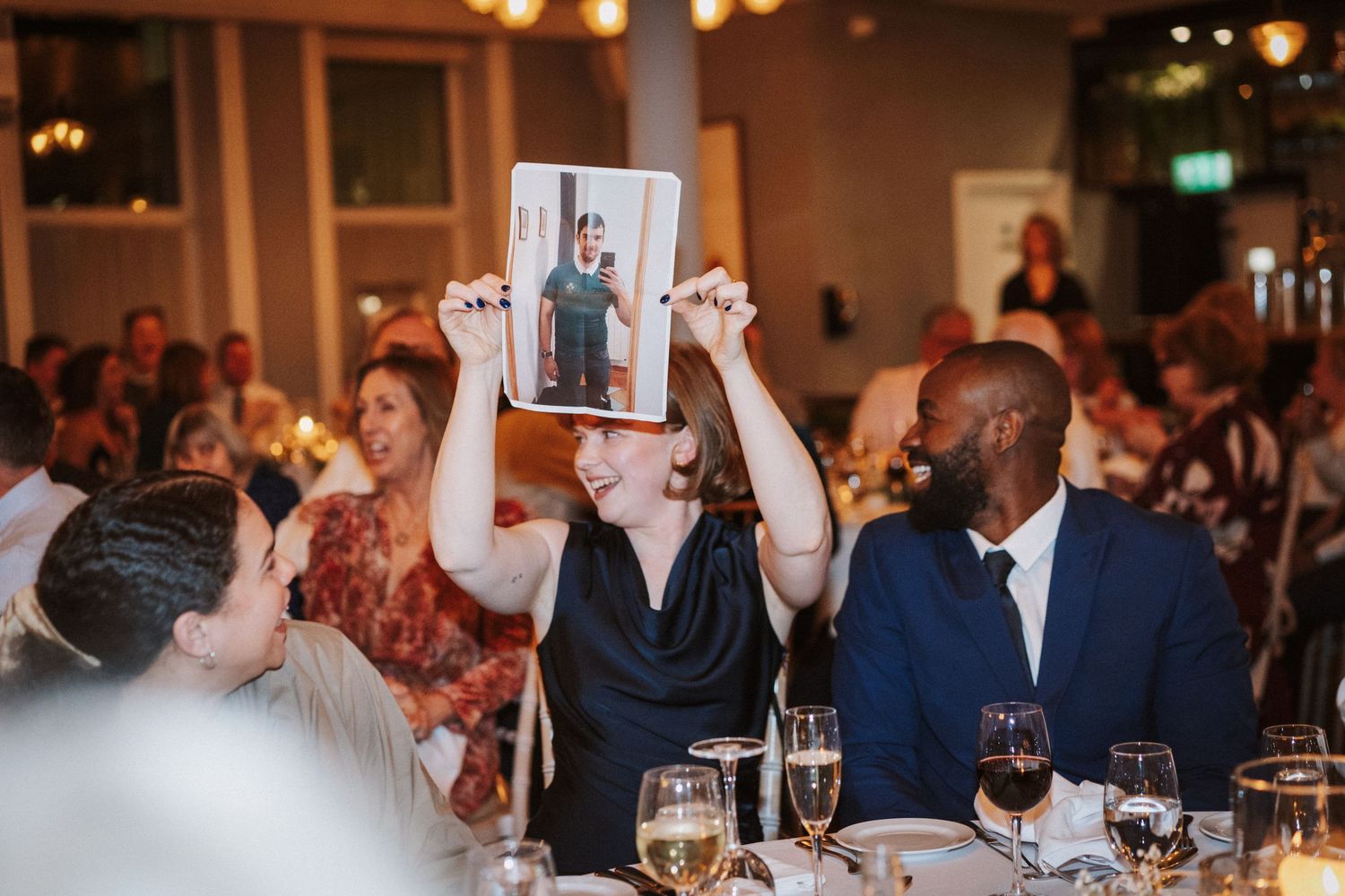 Wedding guests seated at dining tables raise their glasses during a celebratory toast at an evening reception.