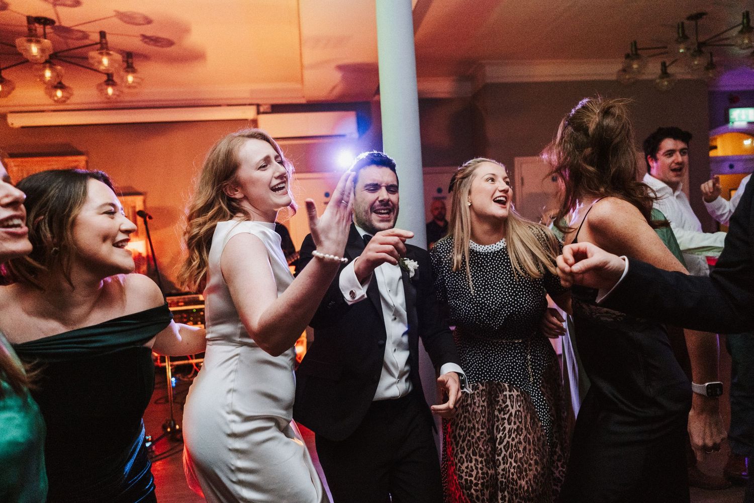 Wedding guests dance and celebrate enthusiastically on a dimly lit dance floor with warm orange lighting.