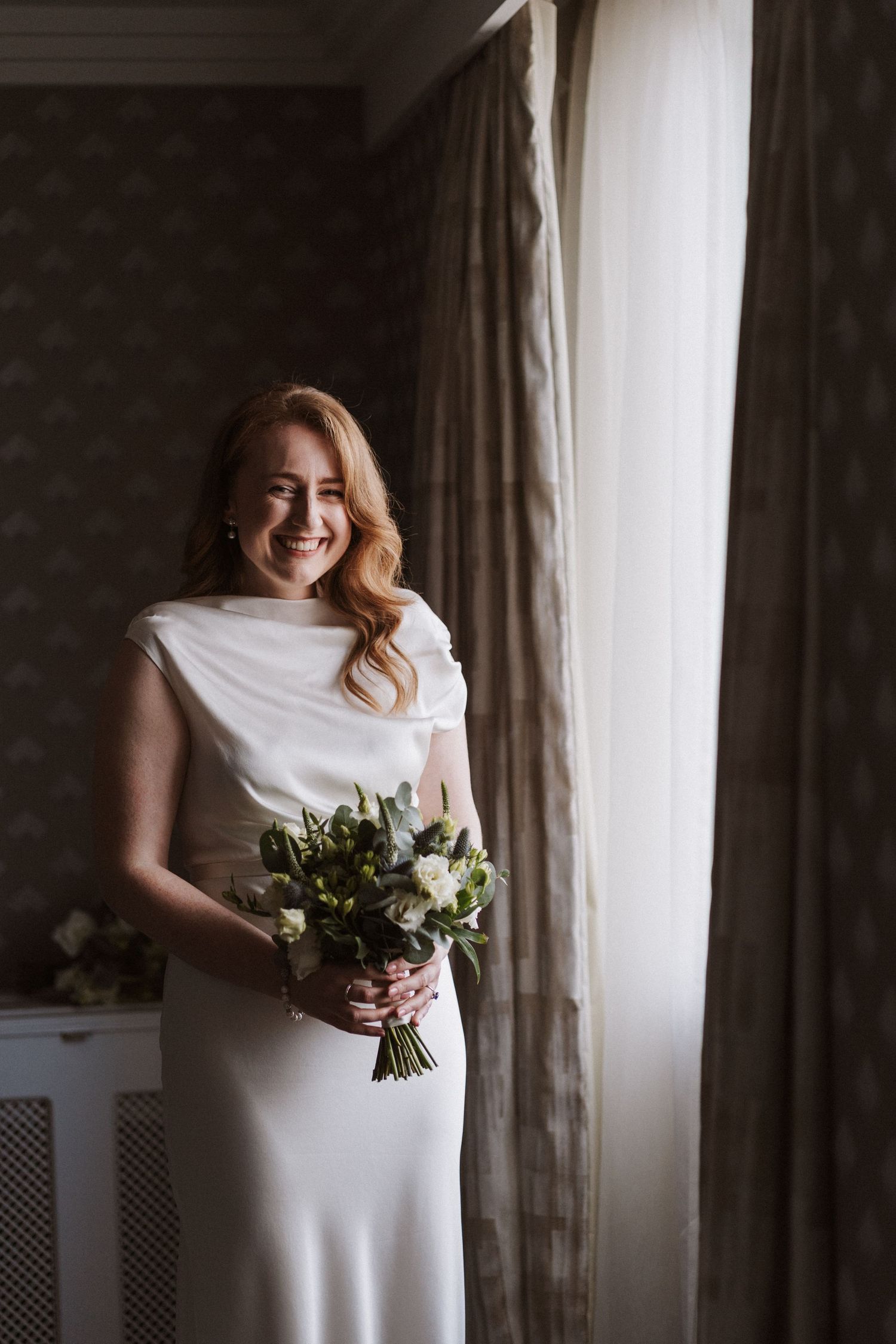 Elegant wedding scene with white bouquet and flowing dress near a sunlit window.