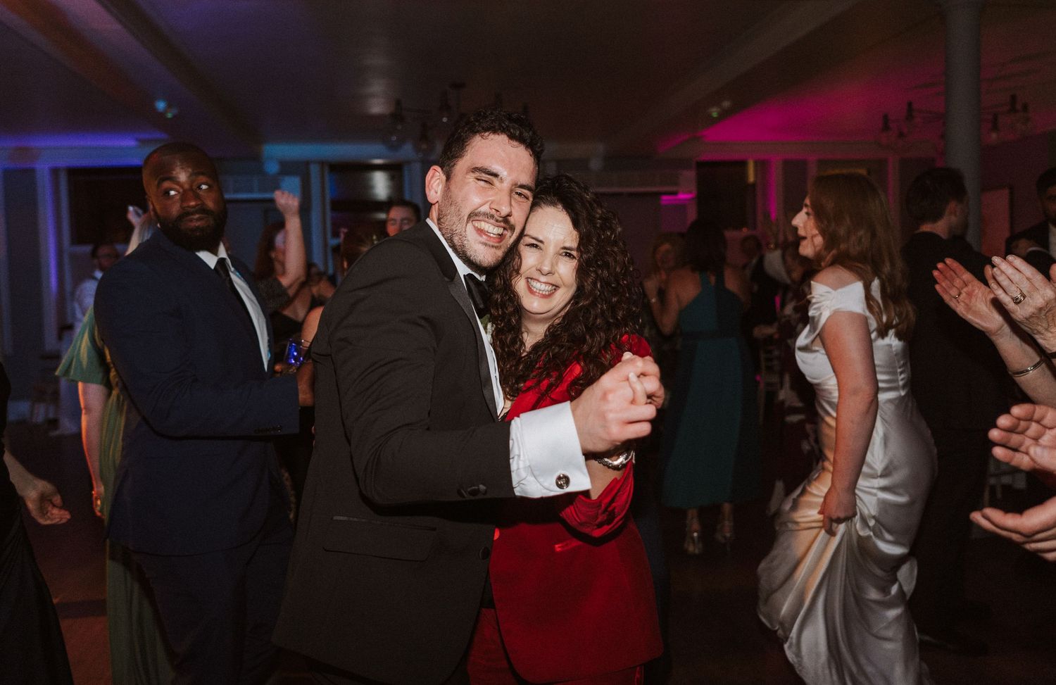 Wedding reception dance floor filled with guests celebrating under colorful mood lighting.