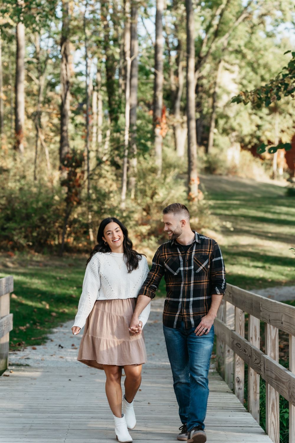 A couple walks together on a bridge in Cuyahoga Falls through a scenic pine forest on a sunny day for a pregnancy announcement photo shoot.