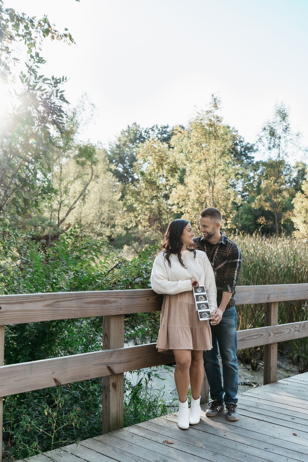 A couple shares a romantic moment on a bridge by lush greenery on a sunny afternoon in Cuyahoga Falls, Ohio for Pregnancy Announcement Pics