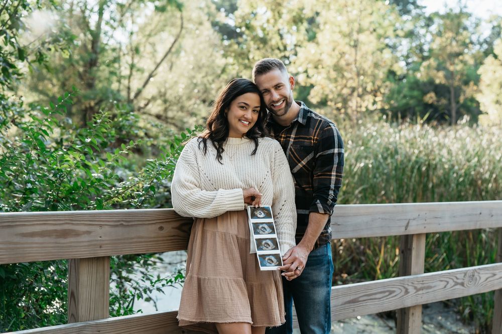A couple stands together on a wooden deck surrounded by lush greenery while holding ultrasound photos at photoshoot in Cuyahoga Falls, Ohio