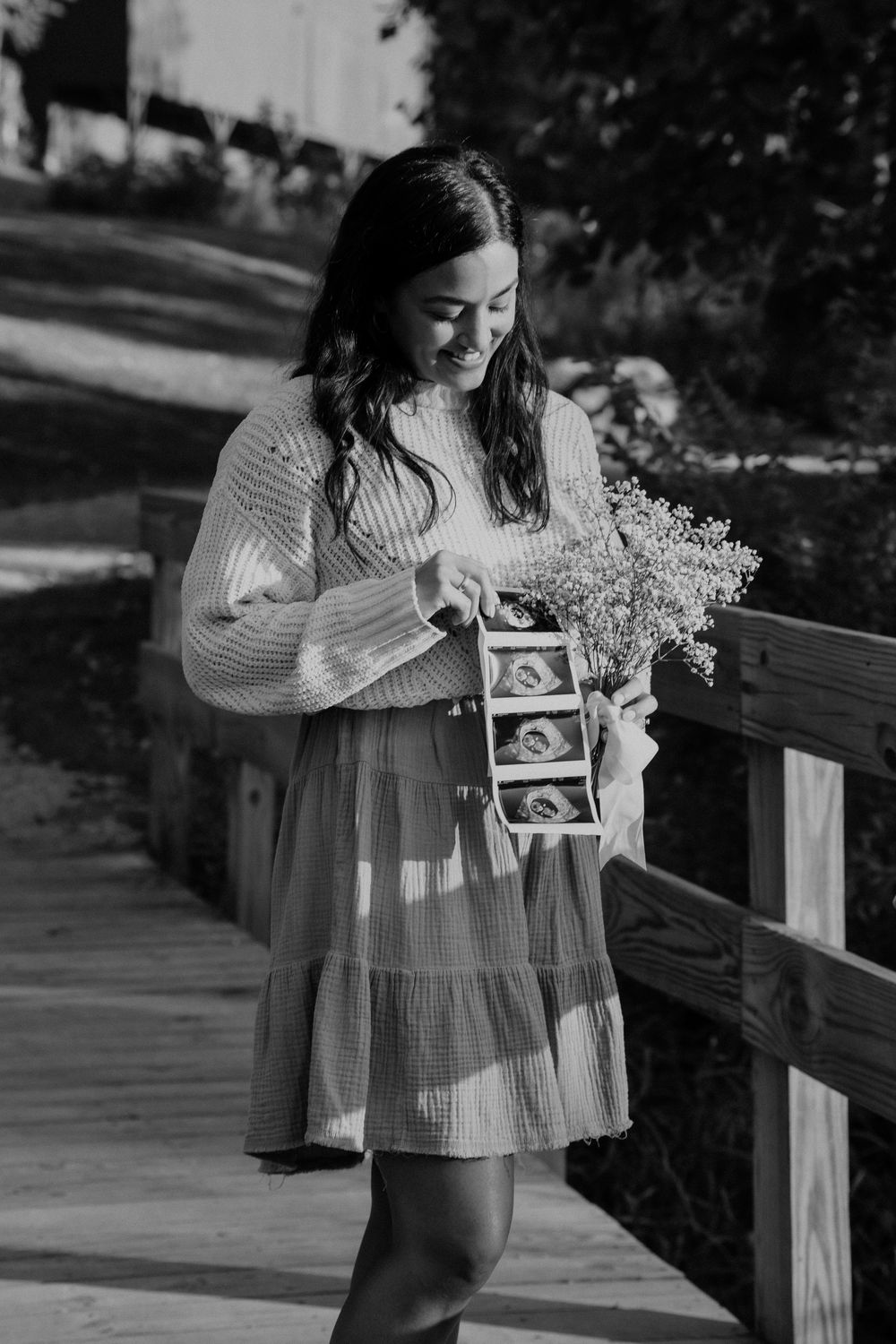 Person in a sweater and flowy dress holds ultrasound photos and a bouquet of baby's breath flowers on a wooden bridge at Mud Brook Greenway