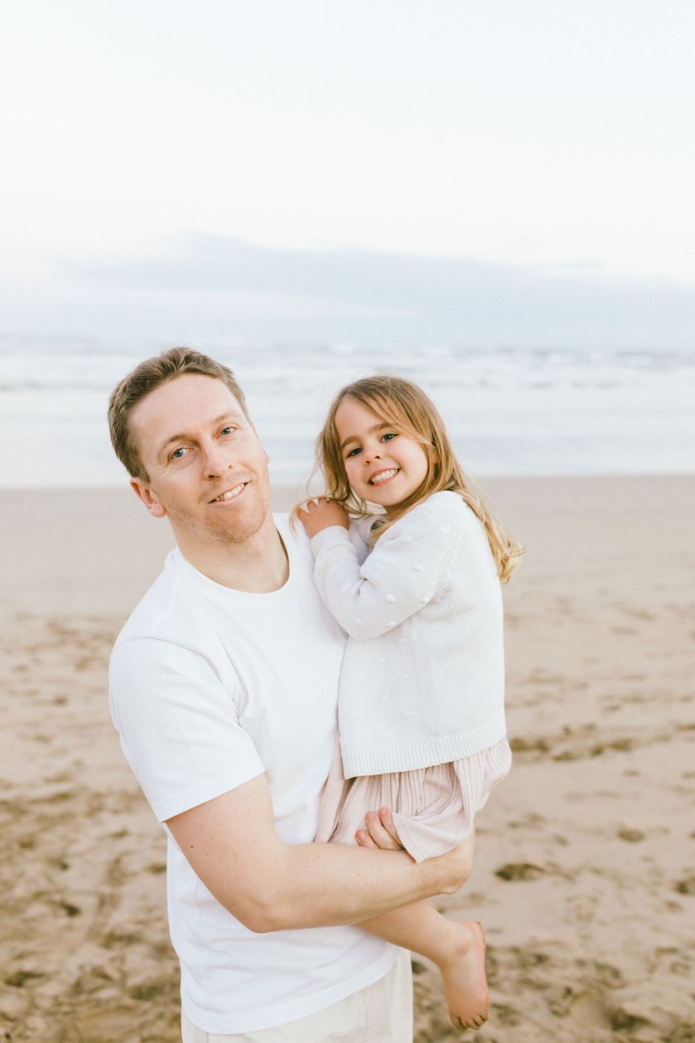 A parent and child in white shirts share joyful moments together on a sandy beach.
