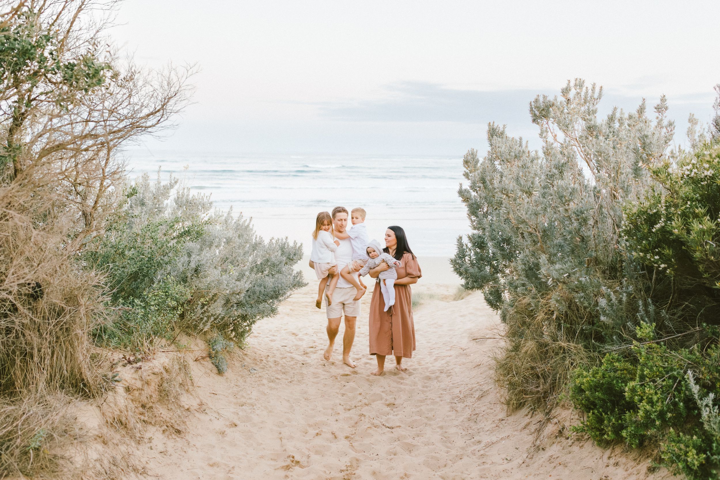 A family walks together on a sandy beach path lined with coastal vegetation and ocean views in the background.