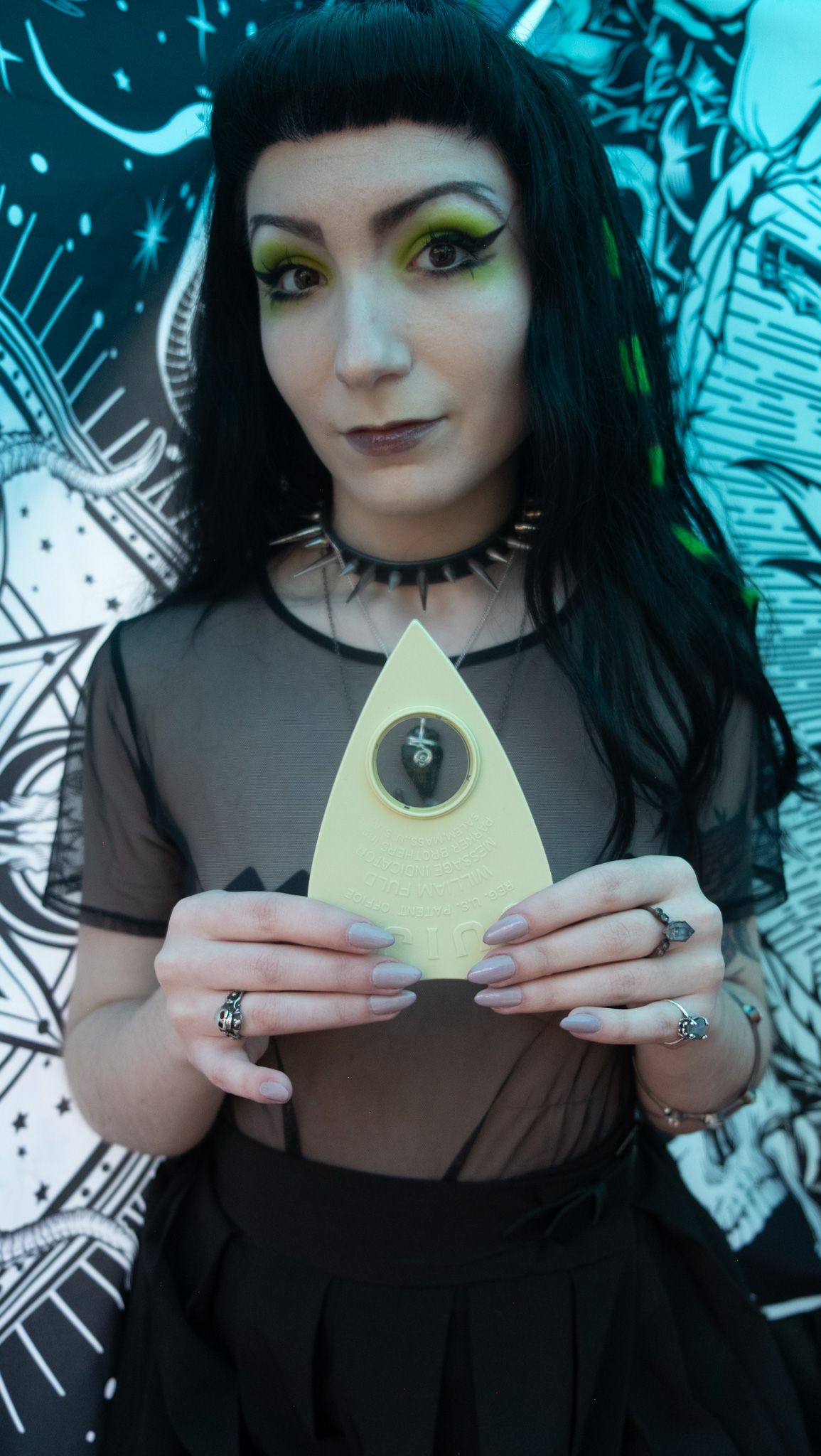 Alternative style model holds planchette prop against blue street art background.