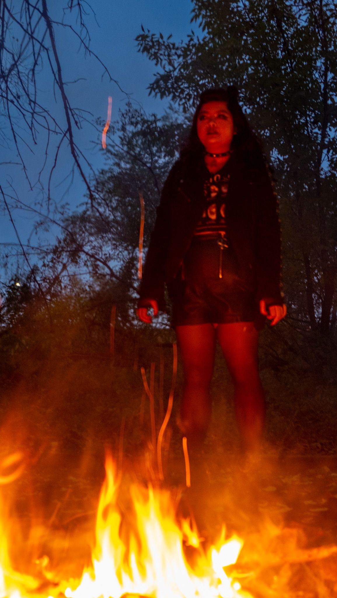 A bright campfire illuminates the dark night sky while casting warm orange light on the surrounding trees and ground.
