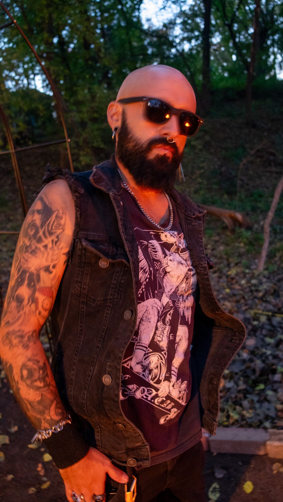 A person in a denim vest with tattoos poses against a wooded background in warm evening light.