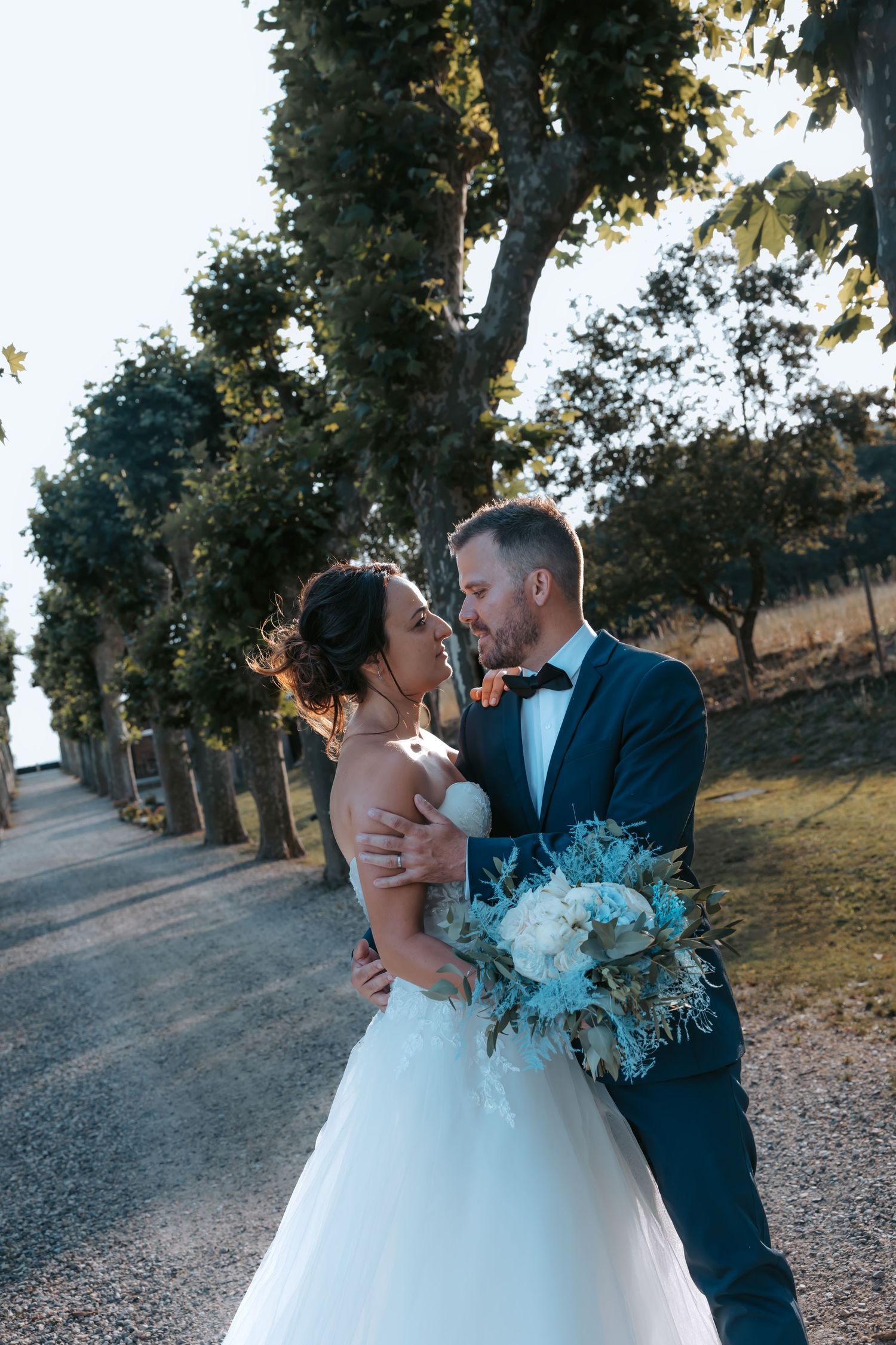 Newlyweds embrace on tree-lined path during outdoor wedding photoshoot.