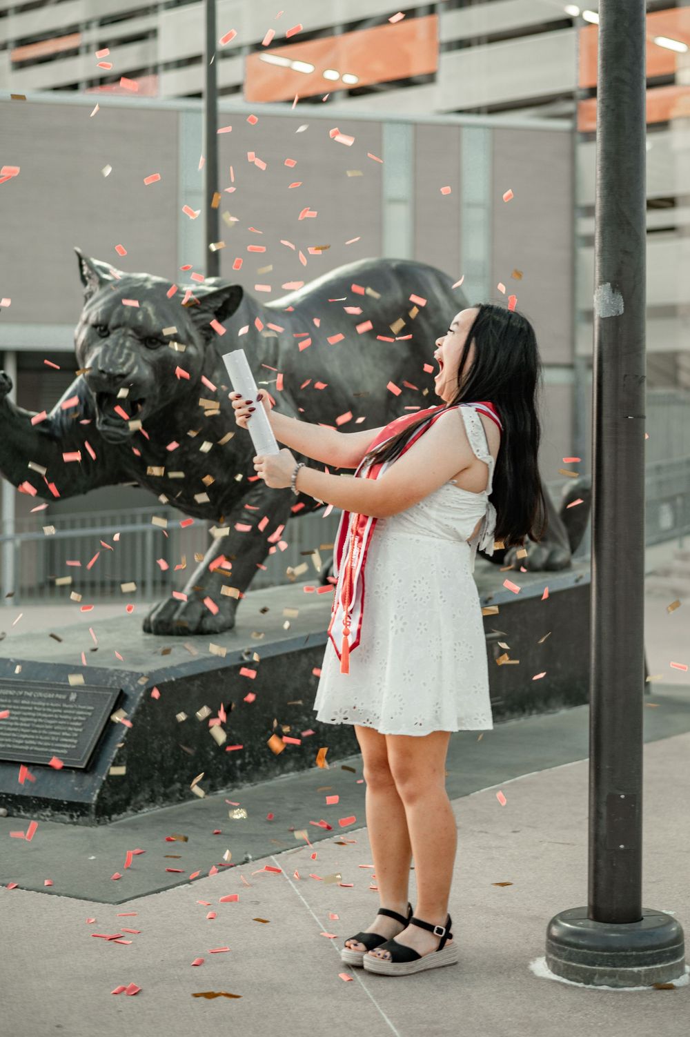Graduate in white dress throwing confetti in front of a cougar statue on campus.