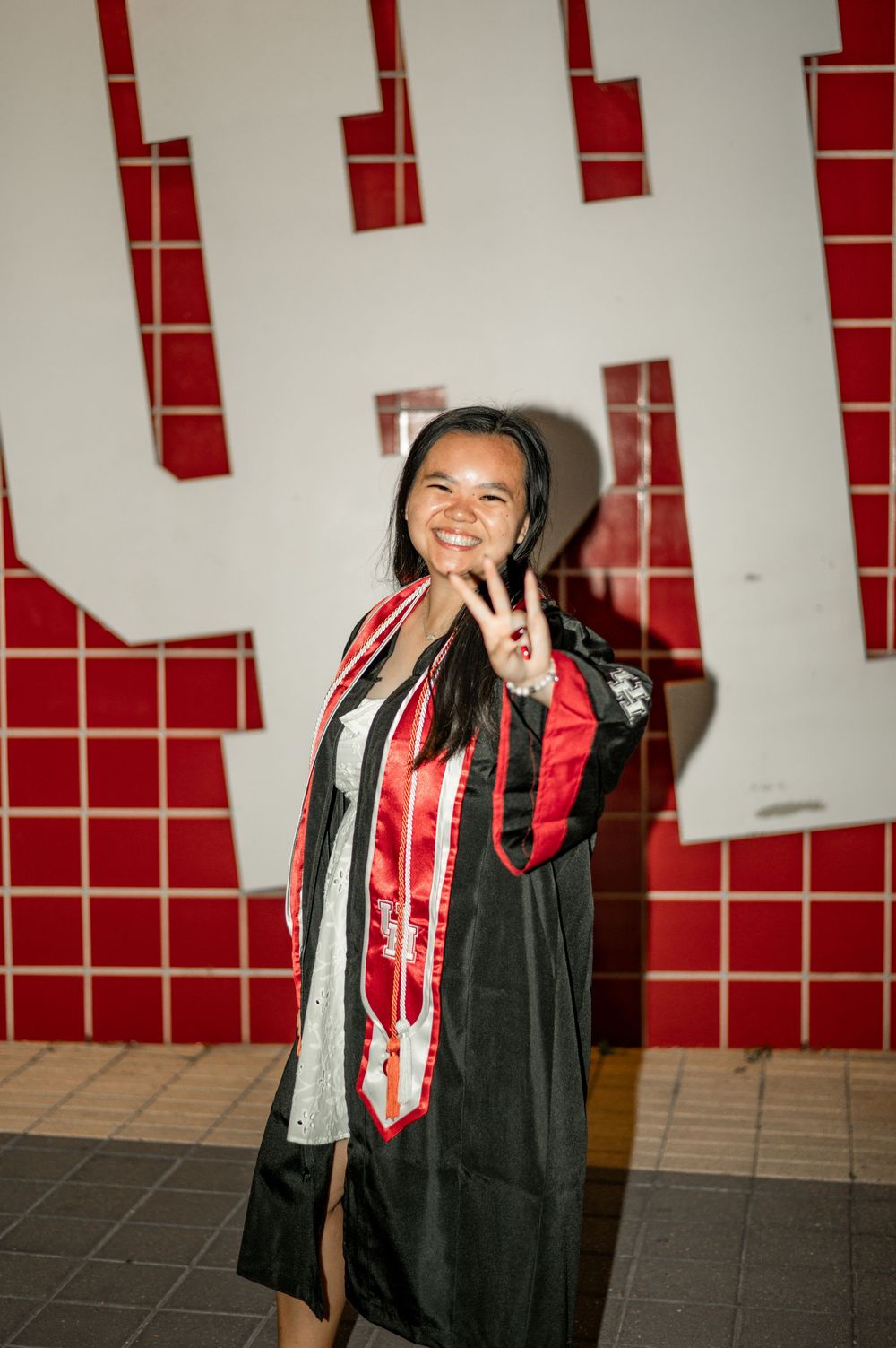 Graduate wearing red and black academic regalia poses against a wall with red decorative elements.