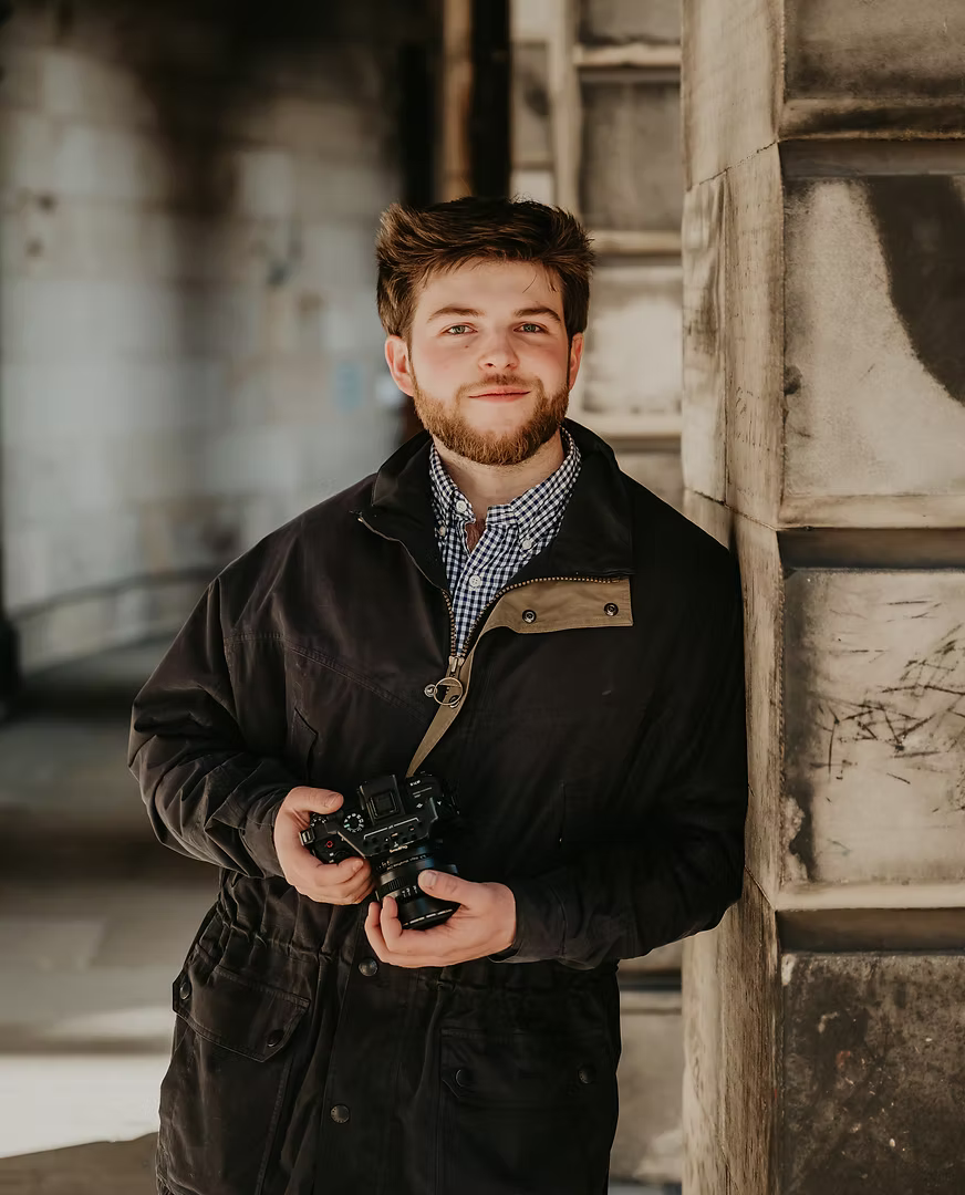 Person in a black coat holds a camera while standing near architectural columns in moody lighting.