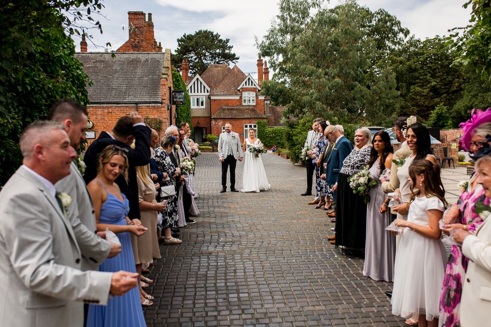 Wedding guests line a cobblestone path outside a red brick building during an outdoor ceremony.