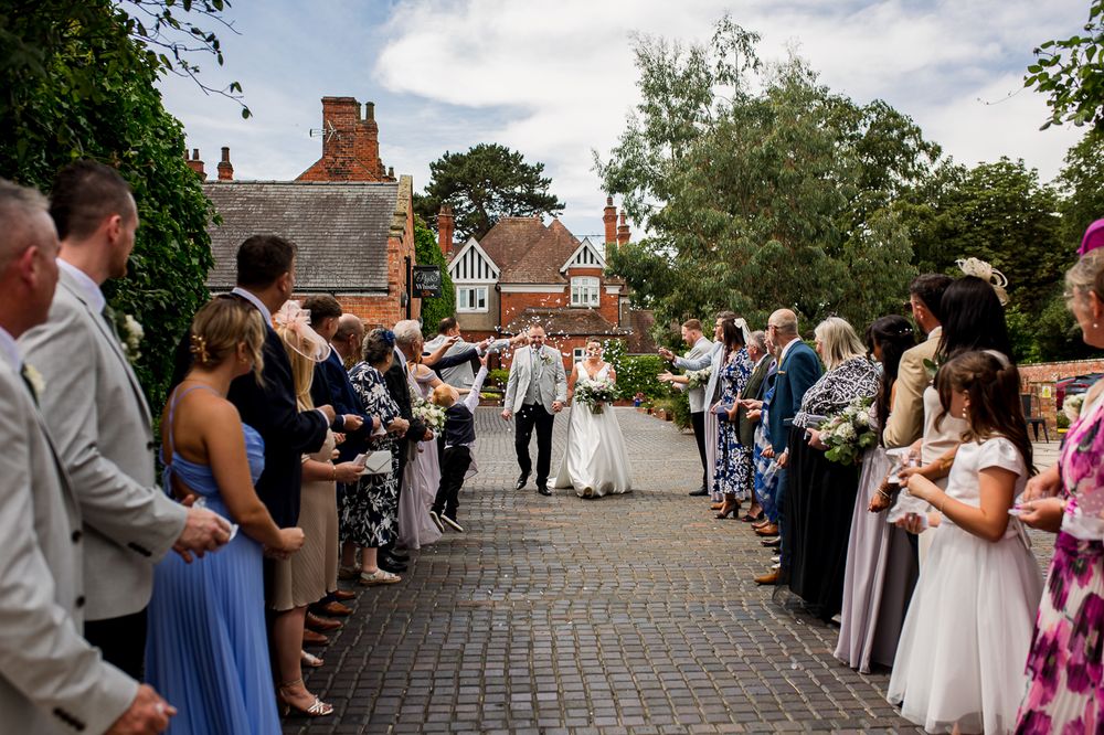 Guests watch wedding ceremony on stone pathway with historic brick building in background.