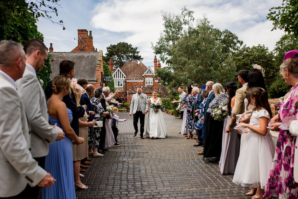 Wedding ceremony guests line stone pathway leading to red brick building entrance.