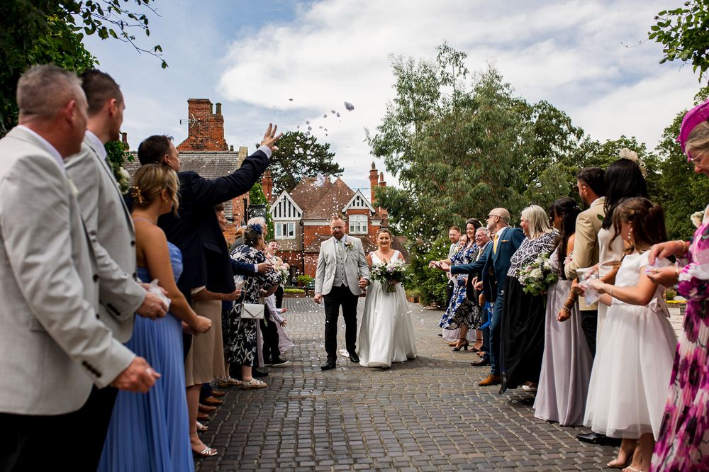 Outdoor wedding ceremony with guests gathered along cobblestone path near historic building.