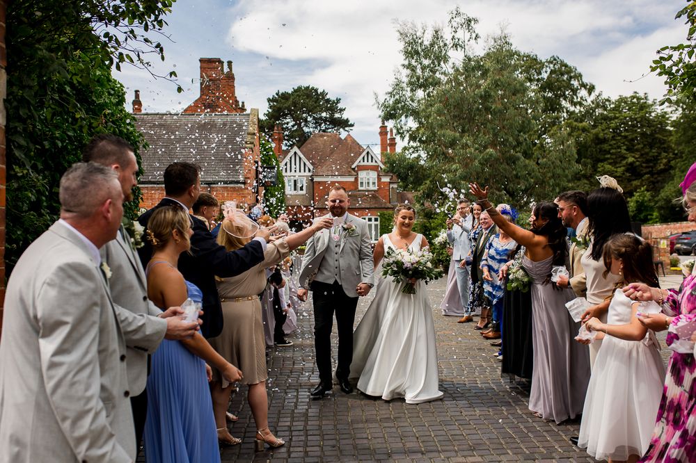 Wedding guests celebrate on stone pathway outside traditional brick building.