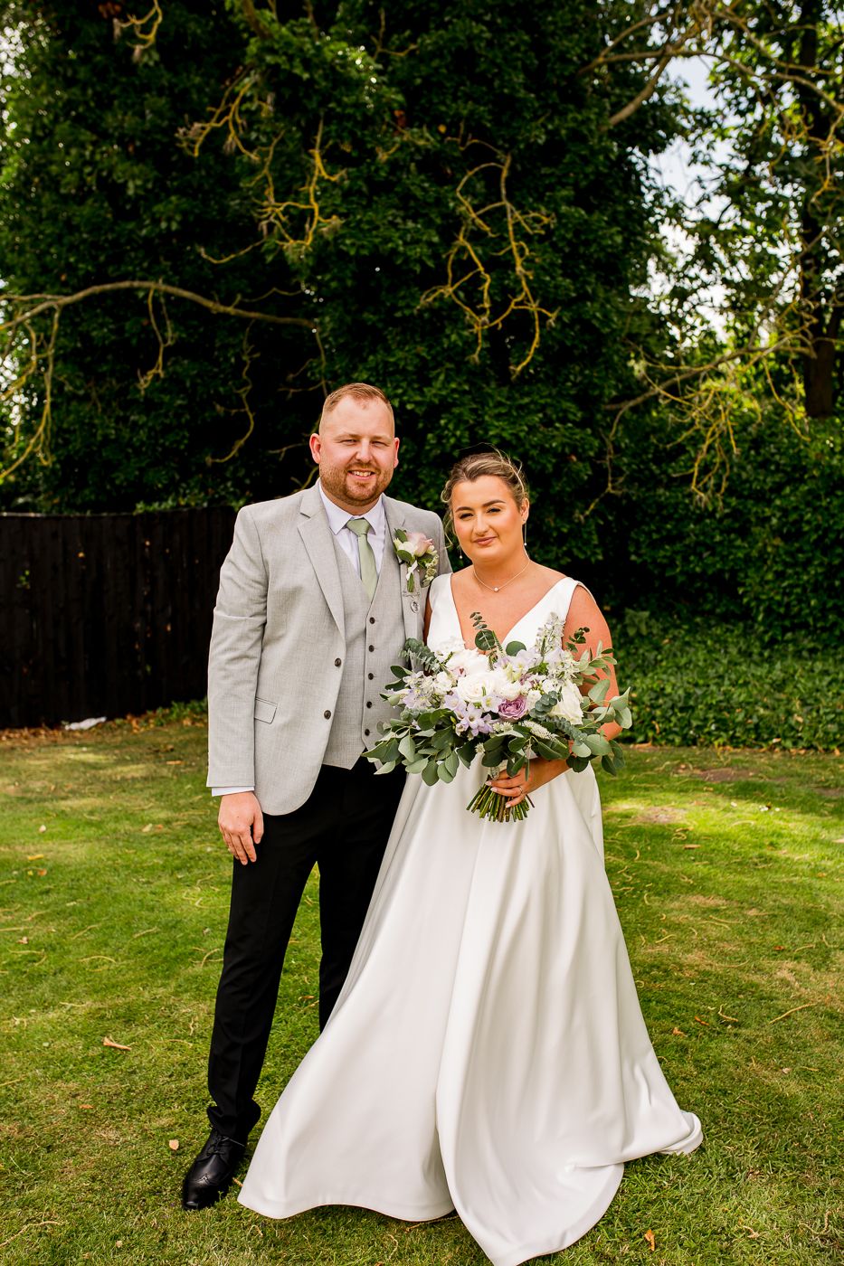 Wedding couple poses on grassy lawn with floral bouquet and greenery in background.