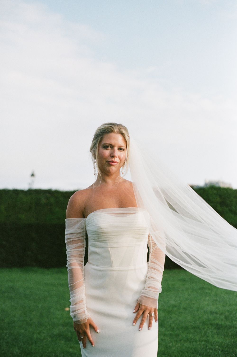 A bride in a simple white wedding dress with long sheer sleeves stands in a grassy field with her veil flowing in the breeze.