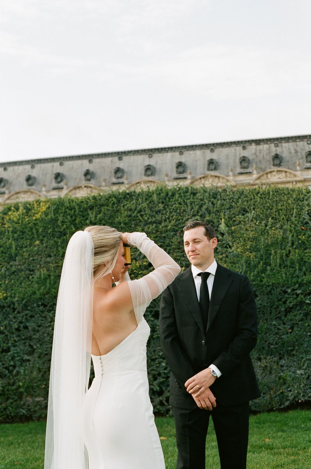 A bride and groom share a candid moment in a garden setting with a stone wall backdrop. Paris editorial elopement photographer.