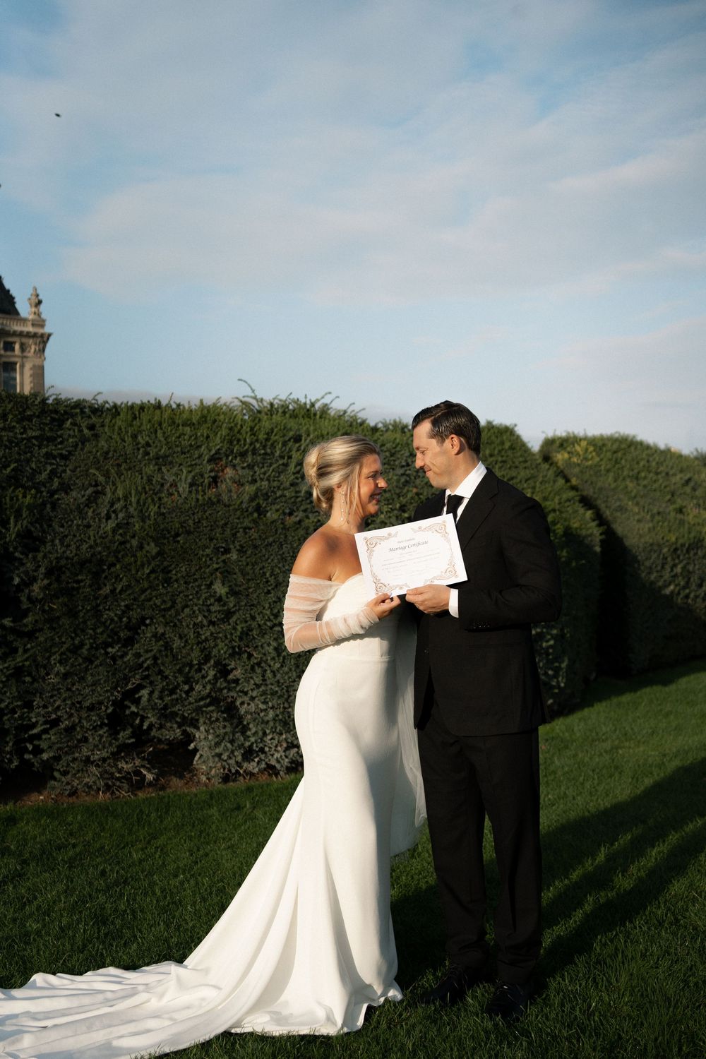 Wedding couple sharing a romantic moment in front of a manicured garden hedge under blue skies. Paris editorial elopement photographer.