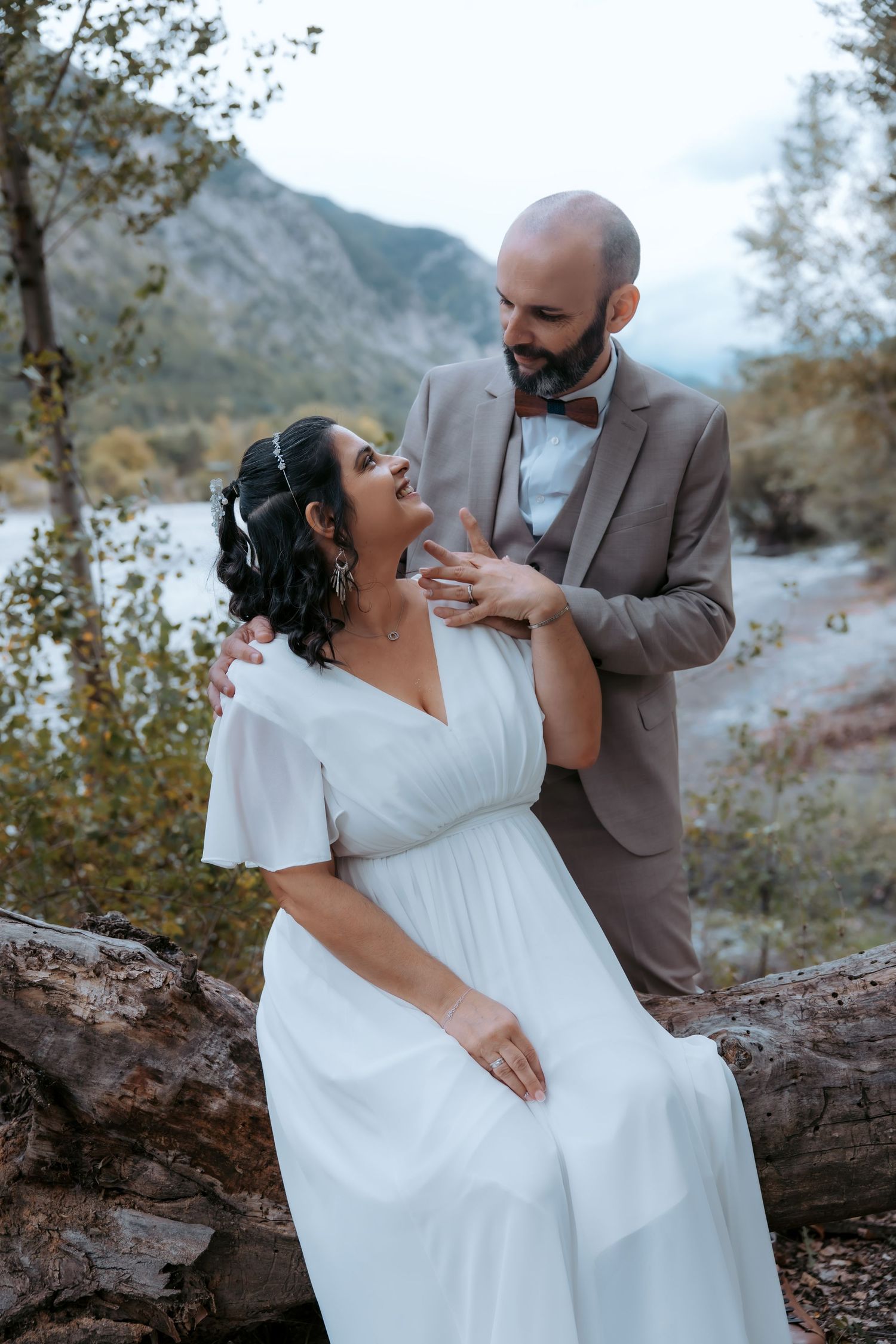 Playful wedding couple sharing intimate moment on fallen tree trunk by mountain lake.