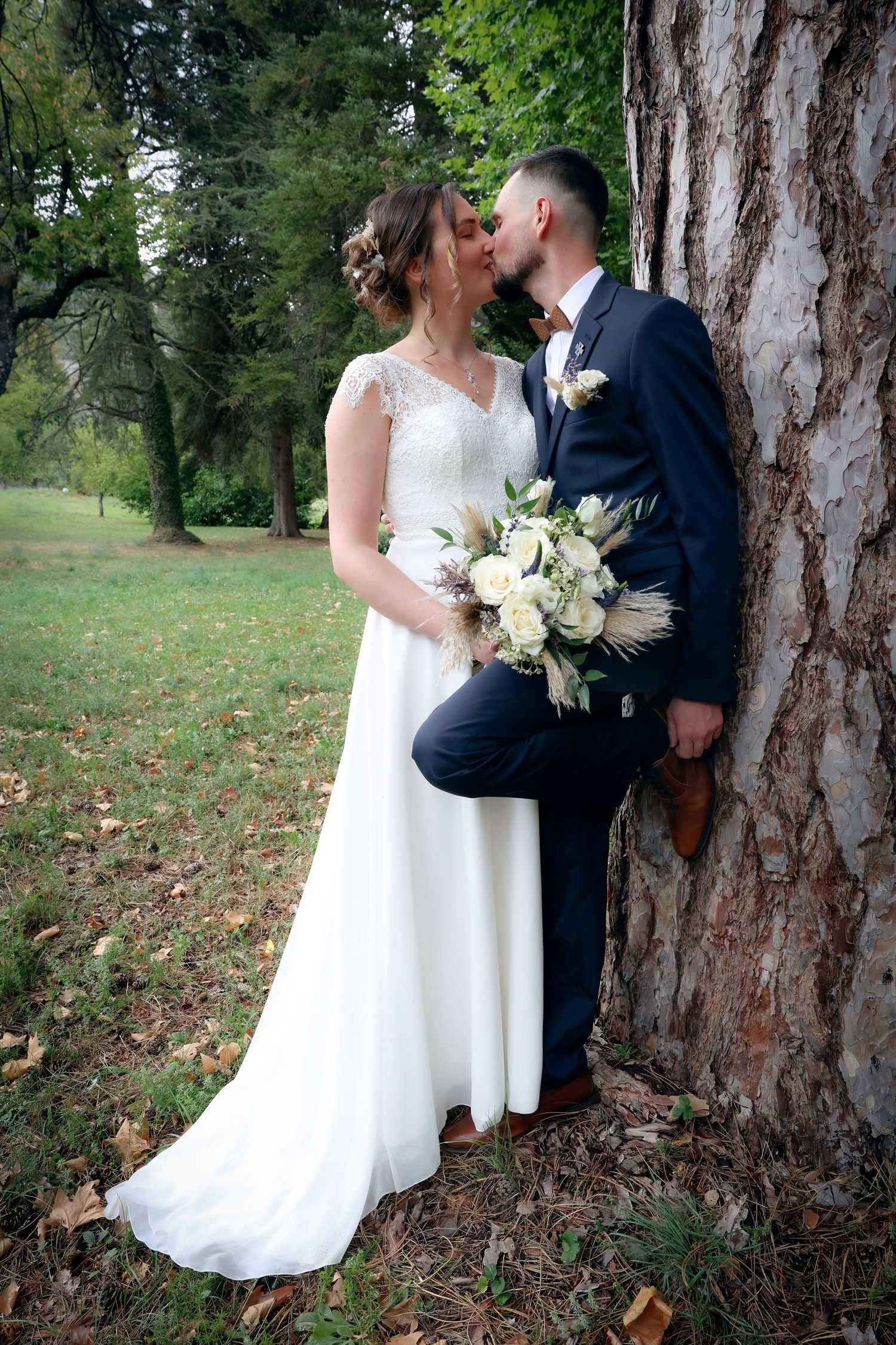 Elegant wedding couple poses against large tree trunk in autumn park setting.