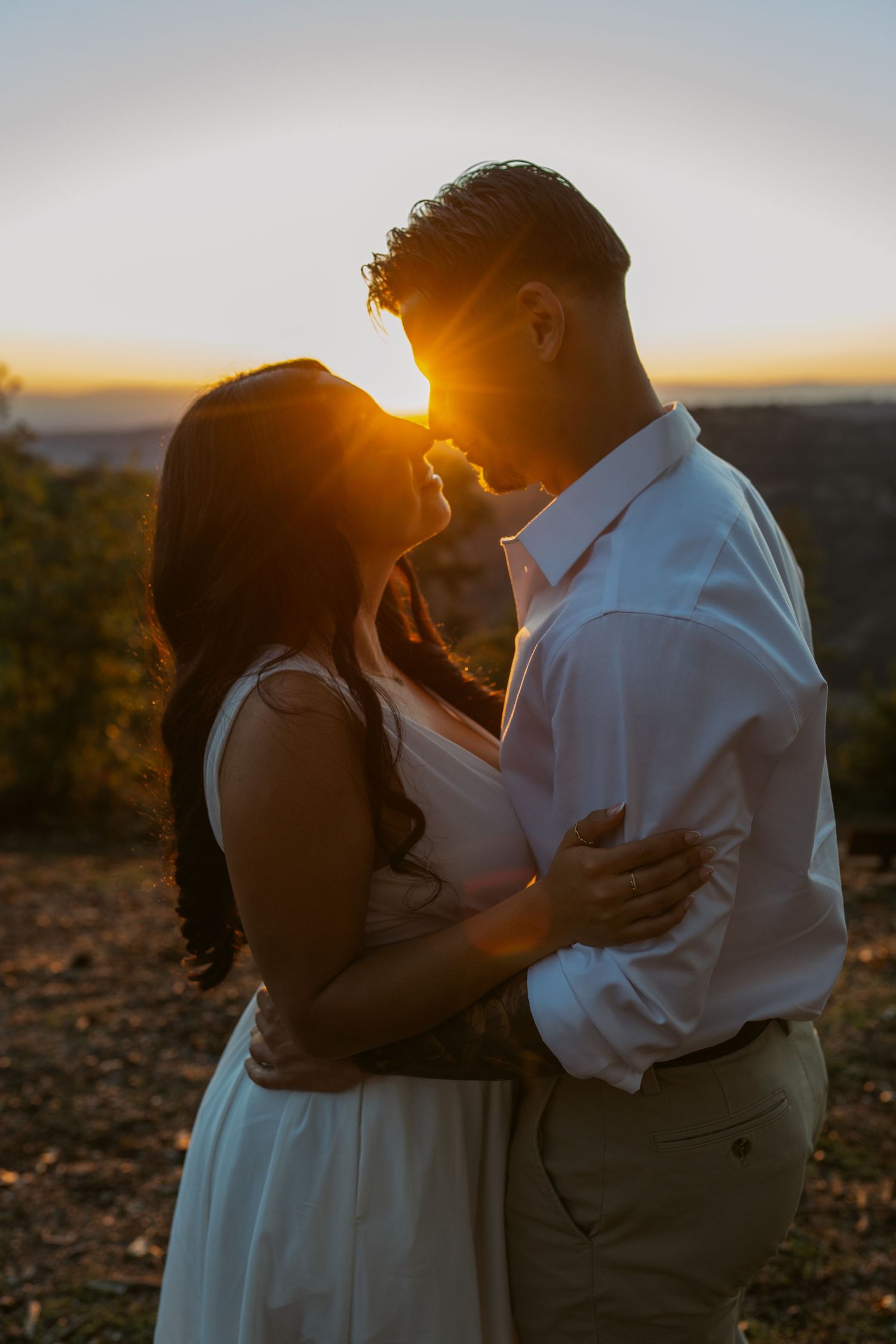 A couple in white clothing sits together on grassy ground during a romantic outdoor photoshoot.