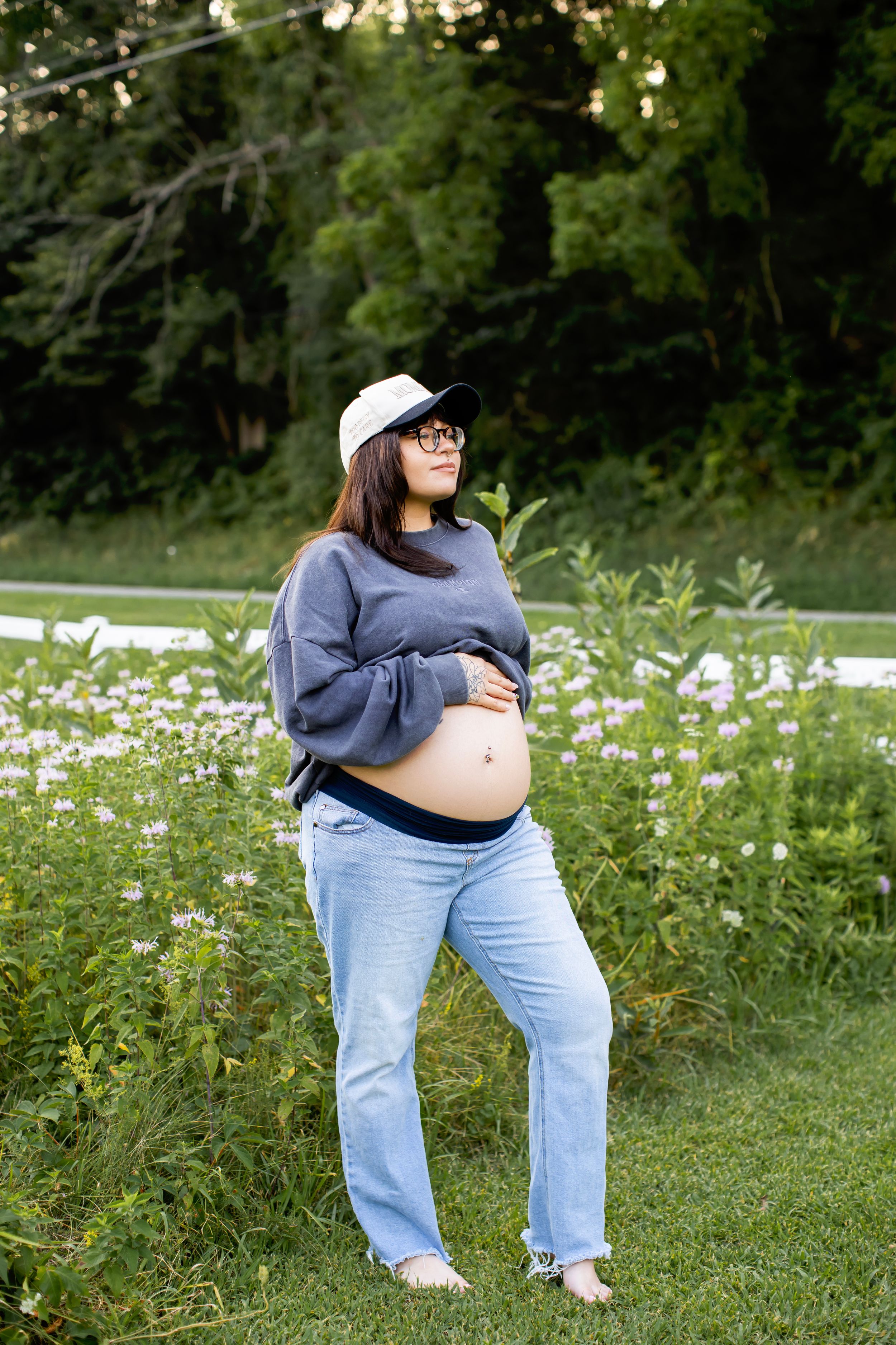 A pregnant person in a blue sweater and jeans sits in a field of wildflowers on a sunny day.